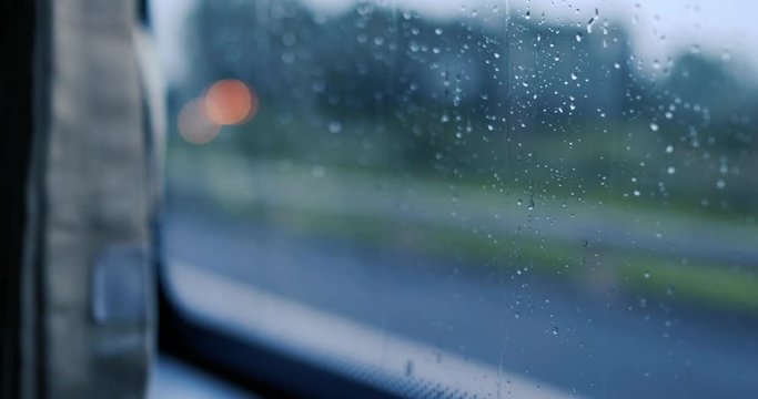 Raindrops on glass car window with road light bokeh. Royalty high-quality free stock footage of rain drops on car glass in rainy season. Closeup of water drop on the glass, car driving in rainy day