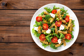 Italian pasta salad with wholegrain fusilli, fresh tomato, cheese, lettuce and broccoli on wooden rustic background. Mediterranean cuisine. Cooking lunch. Healthy diet food. Top view