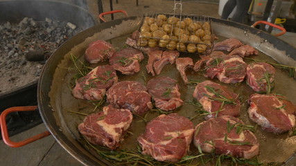 Marinated meat steaks with rosemary sprigs are laid out on a large round frying pan, potatoes for grilling, a chamber along the frying pan
