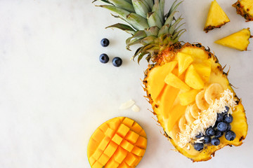 Healthy smoothie bowl in a pineapple with with coconut, bananas, mango and blueberries. Top view on a marble background. Copy space.