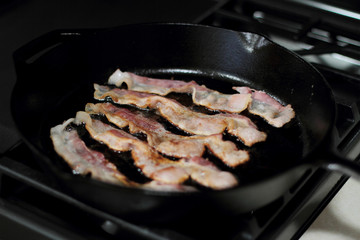Strips of bacon frying in a cast iron pan on the stove.