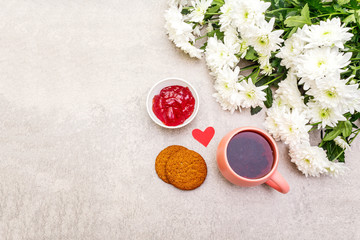 Romantic breakfast concept. A cup of black tea, oatmeal cookies, cherry jelly, bouquet of chrysanthemums. For Valentine's Day or March 8, on a stone background, top view