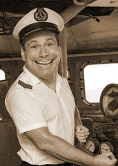 A laughing sailor officer in the uniform is steering the ship with a rudder. The funny captain standing in the wheelhouse of ship and looking to camera, sepia color. © milkovasa