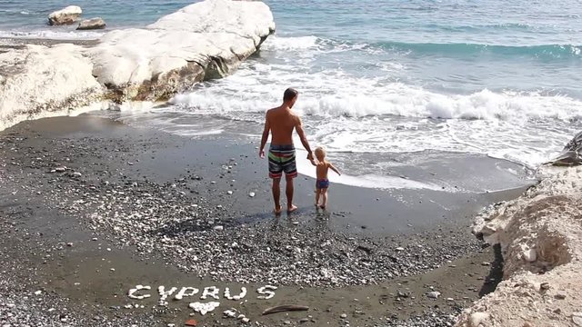 Father and his toddler son play on famous Governor's beach with black sand and white rocks. Inscription CYPRUS from stones at the water's edge.