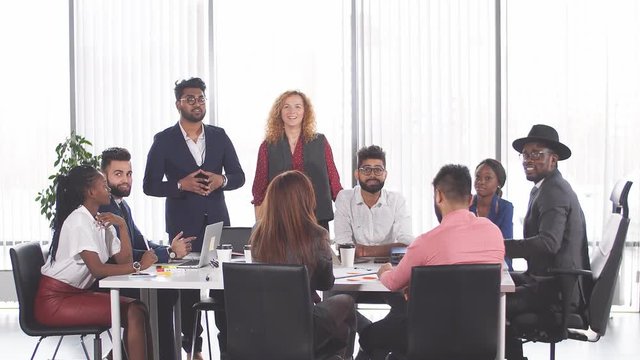Red-haired business woman in cooperation with indian male colleague giving presentation to their interracial coworkers sitting in meeting room