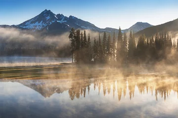 South Sister und Broken Top spiegeln sich bei Sonnenaufgang in der Kaskadenkette in Zentral-Oregon, USA, in einem frühen Morgenlicht über den ruhigen Gewässern des Sparks Lake wider. Morgennebel steigt vom See in die Bäume. © Michal