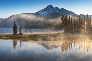 Fototapeta na wymiar South Sister and Broken Top reflect over the calm waters of Sparks Lake at sunrise in the Cascades Range in Central Oregon, USA in an early morning light. Morning mist rises from lake into trees. 