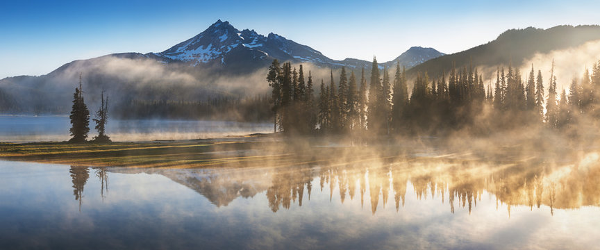 South Sister and Broken Top reflect over the calm waters of Sparks Lake at sunrise in the Cascades Range in Central Oregon, USA in an early morning light. Morning mist rises from lake into trees. 