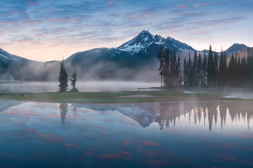 South Sister and Broken Top reflect over the calm waters of Sparks Lake at sunrise in the Cascades Range in Central Oregon, USA in an early morning light. Morning mist rises from lake into trees. 