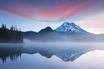South Sister and Broken Top reflect over the calm waters of Sparks Lake at sunrise in the Cascades Range in Central Oregon, USA in an early morning light. Morning mist rises from lake into trees. 