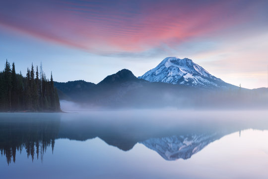 South Sister And Broken Top Reflect Over The Calm Waters Of Sparks Lake At Sunrise In The Cascades Range In Central Oregon, USA In An Early Morning Light. Morning Mist Rises From Lake Into Trees. 