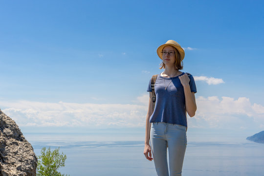 Cute tween tourist girl in hat and backpack standing on cliff top and posing against beautiful landscape of blue sky and Baikal lake. Hiking, travelling and summer vacation concept