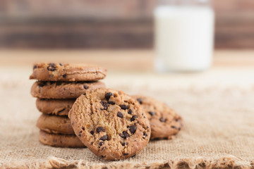A stack of chocolate chip cookies and bottles of milk on sackcloth on wood table.