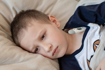 Pre-school sick boy in pyjama lying in bed with a digital thermometer.