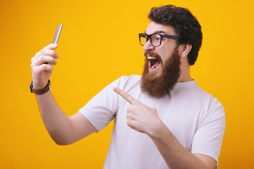 Guy eyeglasses cheerful pointing at smartphone. Bearded man happy user interact application for...