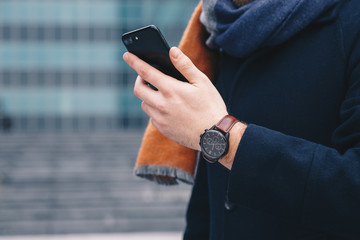 Detail shot of a male hand using a mobile phone and wearing an analog hand watch, in an urban surroundings.