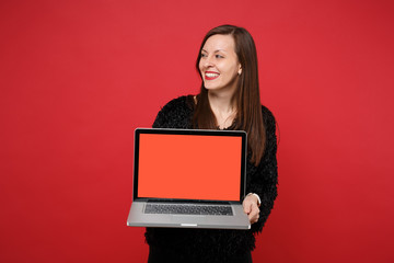 Naklejka premium Joyful young woman in black fur sweater looking aside hold laptop pc computer with blank empty screen isolated on bright red background. People sincere emotions, lifestyle concept. Mock up copy space.