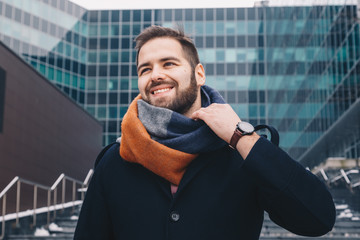 Portrait of a successful and gashionable man posing in front of a corporate modern day building, smiling and looking up while fixing his scarf.