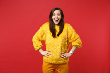 Excited young woman in yellow fur sweater with opened mouth standing with arms akimbo isolated on bright red wall background in studio. People sincere emotions, lifestyle concept. Mock up copy space.