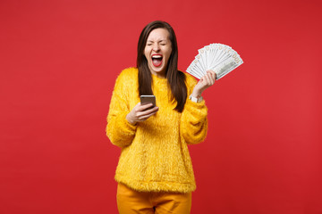 Screaming young woman with closed eyes holding mobile phone, fan of money in dollar banknotes, cash money isolated on red background. People sincere emotions, lifestyle concept. Mock up copy space.