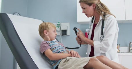 Female pediatrician checking ears of young boy in clinic exam room. Woman doctor giving exam using otoscope to child in pediatrician office. Slow motion 4k - Powered by Adobe