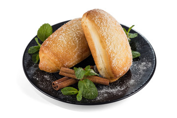 Two buns of pastry, decorated with fresh mint leaves and cinnamon sticks, lie on a black plate, isolated on a white background with shadows.