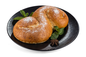 Fresh butter bun in the form of Latin letter S sprinkled with powdered sugar and decorated with fresh mint leaves, lying on a black plate isolated on a white background with shadows.