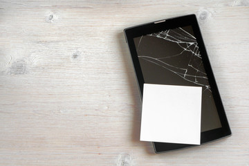 Broken tablet computer with cracked screen, with clean sheet of paper on wooden background. Copy...