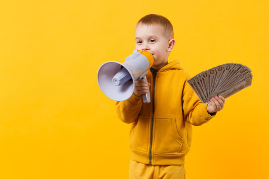 Little happy kid boy in yellow clothes hold fan of money in dollar banknotes, megaphone isolated on orange wall background, children studio portrait. Childhood lifestyle concept. Mock up copy space.