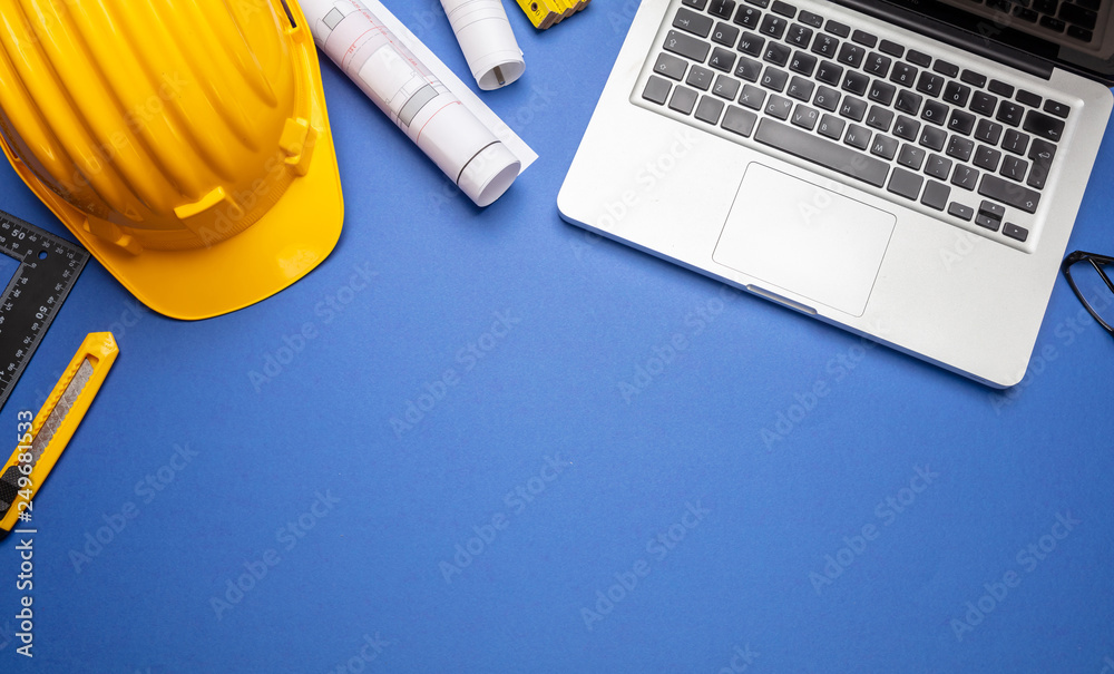 Wall mural project blueprints, yellow hard hat and computer laptop on blue color, top view