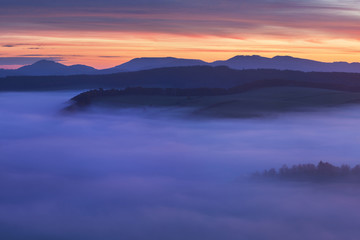 Naklejka na ściany i meble Sunset Over Misty Landscape Scenic View Of Foggy Morning Sky With Rising Sun Above dreamy Forest. Mountain range with visible silhouettes through the morning colorful fog Beautiful background concept