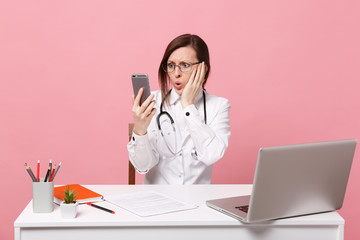 Female doctor sit at desk work on computer with medical document hold cellphone in hospital isolated on pastel pink background. Woman in medical gown glasses stethoscope. Healthcare medicine concept.