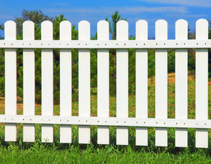White wooden fence on green grass against of the trees and blue sky.