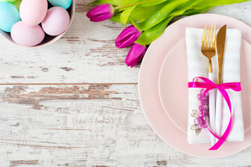 Easter table setting, flatlay with pastel colored Easter eggs, event decoration. Pink dishes and a gold fork and a spoon on a light rustic wooden background. Beautiful purple spring tulip flowers