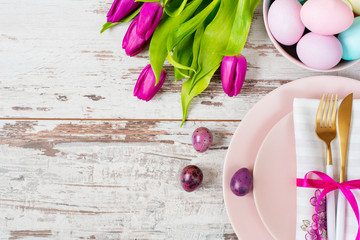 Easter table setting, flatlay with pastel colored Easter eggs, event decoration. Pink dishes and a gold fork and a spoon on a light rustic wooden background. Beautiful purple spring tulip flowers