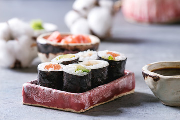 Sushi rolls set in pink ceramic serving plate with bowls of soy sauce and pickled ginger, cotton flowers on light blue table. Japan menu. Close up