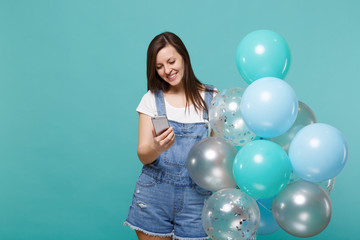 Pretty young woman in denim clothes looking on mobile phone in hand celebrating, hold colorful air balloons isolated on blue turquoise wall background. Birthday holiday party, people emotions concept.