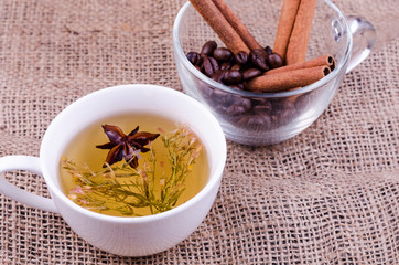 A cup of green tea with flowers and anise against a cup of coffee grains and cinnamon