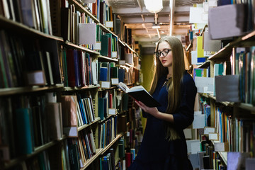 Beautiful young woman wearing glasses in the library among the bookshelves holding a book in her hands and reading, studying.