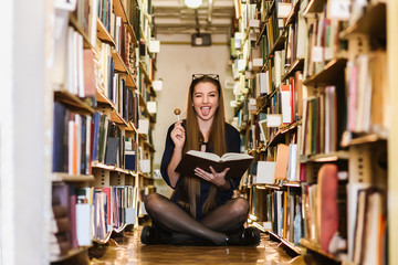 Beautiful young woman wearing glasses in the library among the bookshelves holding a book and candy  in her hands and reading, studying.