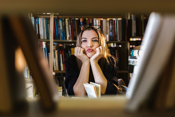 Beautiful young woman wearing glasses in the library among the bookshelves holding glasses and looking at the lens, reading, studying.