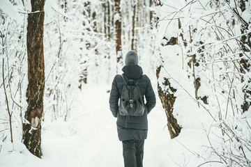 a back shot of a Beautiful youngwoman dressed in a black winter jacket with leather backpack walks in the snowy forest wood .landscape background behind