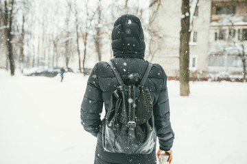 Fototapeta na wymiar a back shot of a Beautiful youngwoman dressed in a black winter jacket with leather backpack walks in the snowy City with the thermos flask in the hand