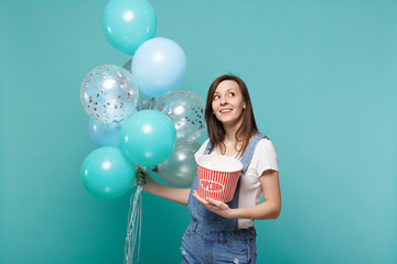 Fototapeta premium Pensive young woman in denim clothes looking up holding bucket of popcorn celebrating with colorful air balloons isolated on blue turquoise background. Birthday holiday party, people emotions concept.