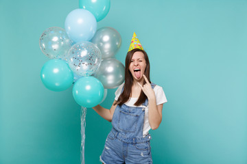 Fototapeta premium Crazy girl in birthday hat showing tongue horns up heavy metal rock sign celebrating, hold colorful air balloons isolated on blue turquoise background. Birthday holiday party, people emotions concept.