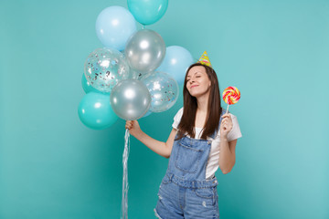 Relaxed woman in birthday hat keeping eyes closed holding round lollipop, celebrating with colorful air balloons isolated on blue turquoise background. Birthday holiday party, people emotions concept.