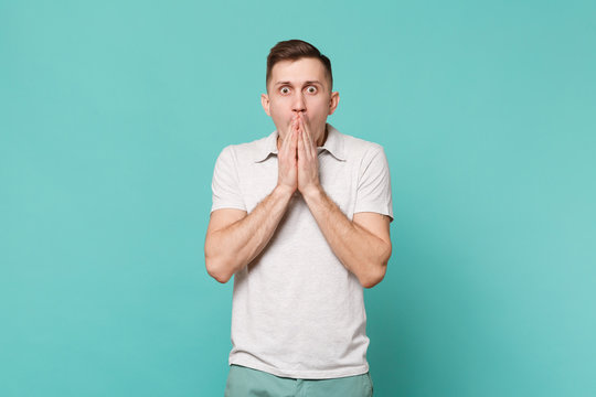 Portrait of shocked young man in casual clothes standing covering mouth with palms isolated on blue turquoise wall background in studio. People sincere emotions, lifestyle concept. Mock up copy space.