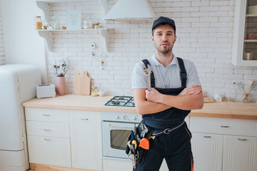 Plumber in kitchen. Professional look on camera and pose. Hands crossed. Tools on belt aside.