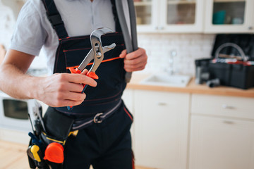 Cut view of man in working uniform hold wrench. He stand in kitchen. Daylight. Tools on belt. Toolbox on desk.