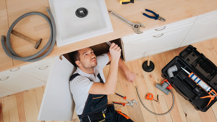 Young plumber look under sink in kitchen. He put new hose. Tools with opened box on floor. Hose and...
